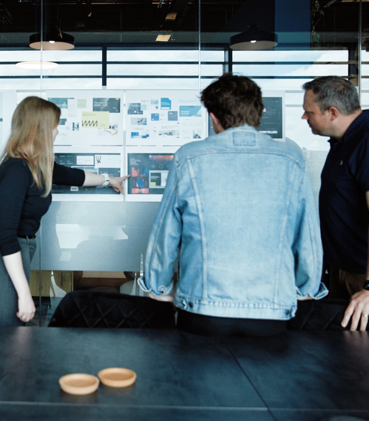Over-the-shoulder view of a branding critique session, with team members reviewing visuals pinned to the wall.